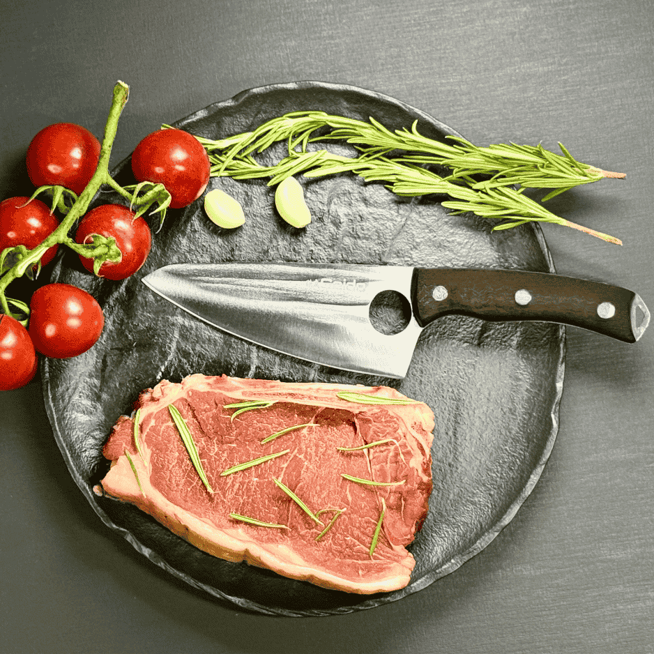 Vachelli Akamori stainless steel chef knife displayed with raw steak, rosemary, garlic, and tomatoes.