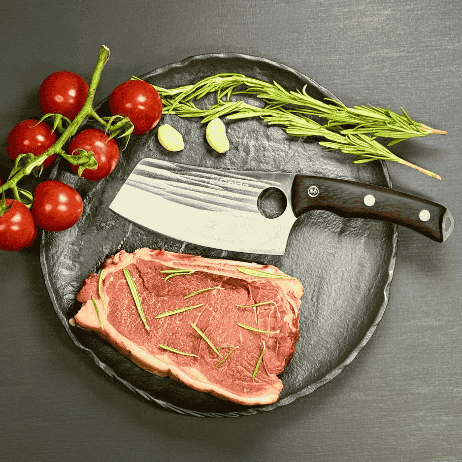 Vachelli Akamori stainless steel cleaver on plate with raw steak, rosemary, garlic, and tomatoes.