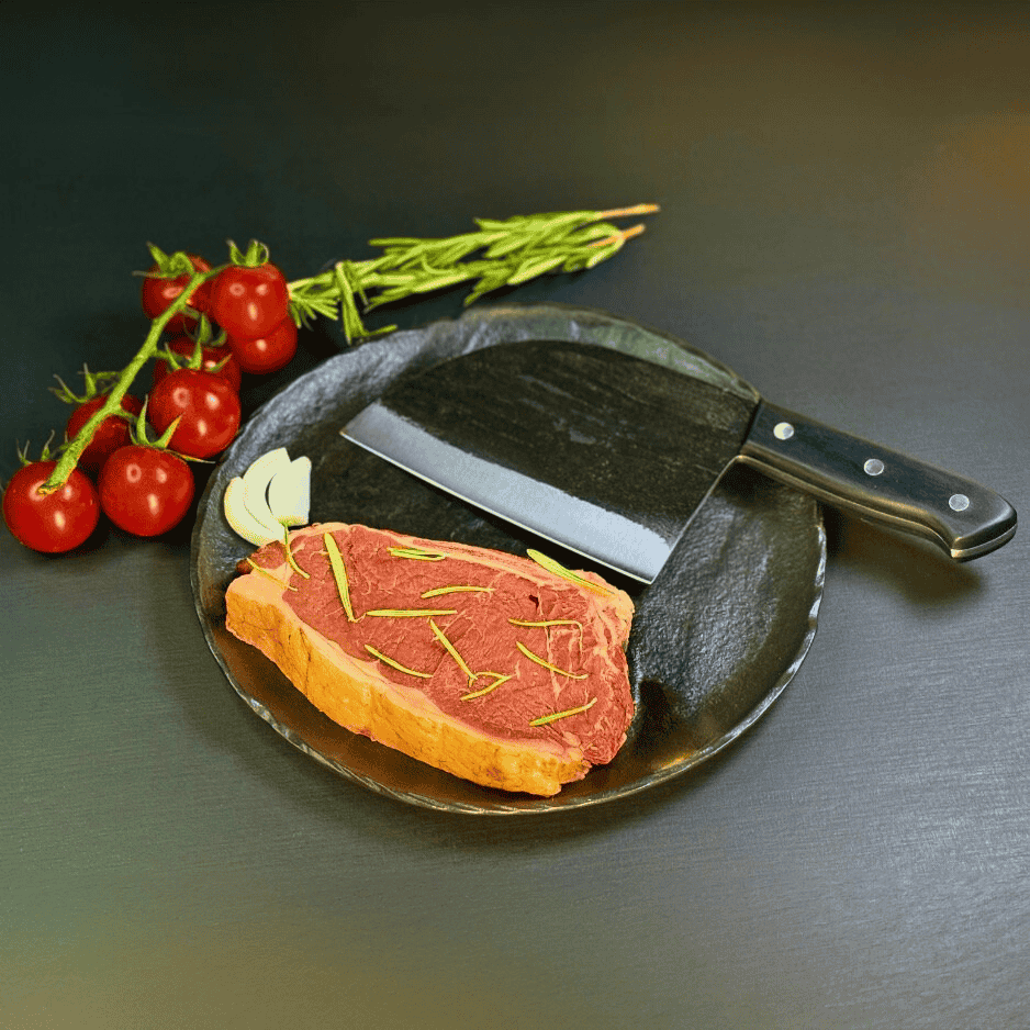 Vachelli Dravok forged carbon steel butcher knife displayed with raw steak, rosemary, onions, and tomatoes.