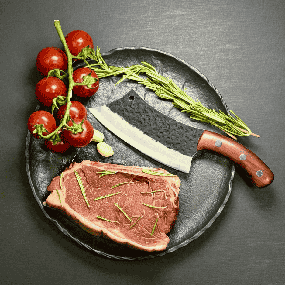 Vachelli Orvian forged carbon steel skinner knife on plate with raw steak, rosemary, garlic, and tomatoes.