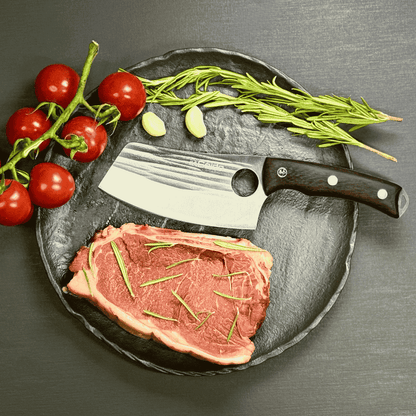 Vachelli Akamori stainless steel cleaver on plate with raw steak, rosemary, garlic, and tomatoes.