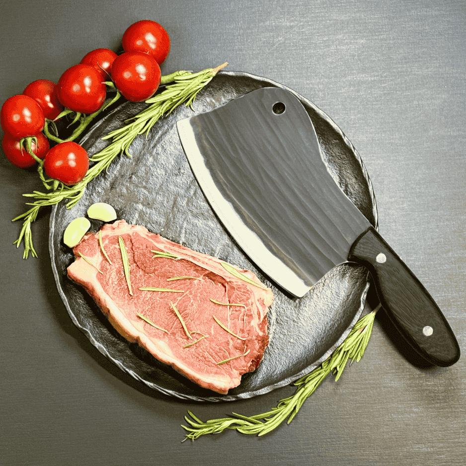 Vachelli Torvani forged carbon steel cleaver on plate with raw steak, rosemary, garlic, and tomatoes.
