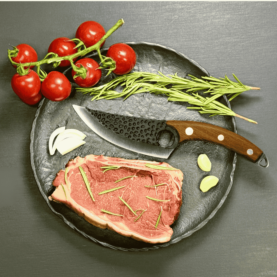 Vachelli Velari steak knife with hammered carbon steel blade and wooden handle, displayed on a black plate with raw steak, garlic, rosemary, and vine tomatoes.